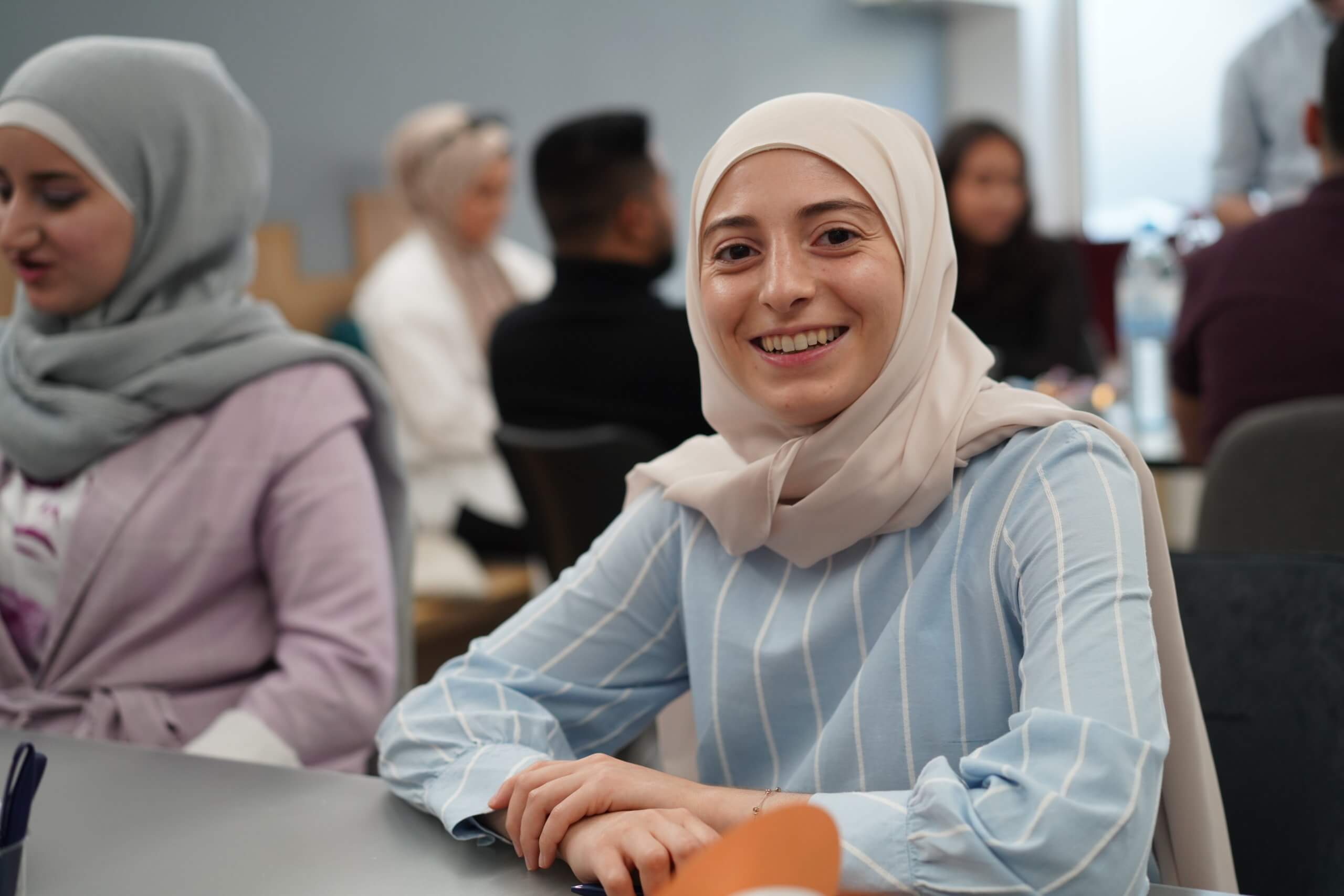 Eine junge Frau mit Hijab lächelt in die Kamera und sitzt in einem Workshopraum mit anderen Teilnehmer:innen.