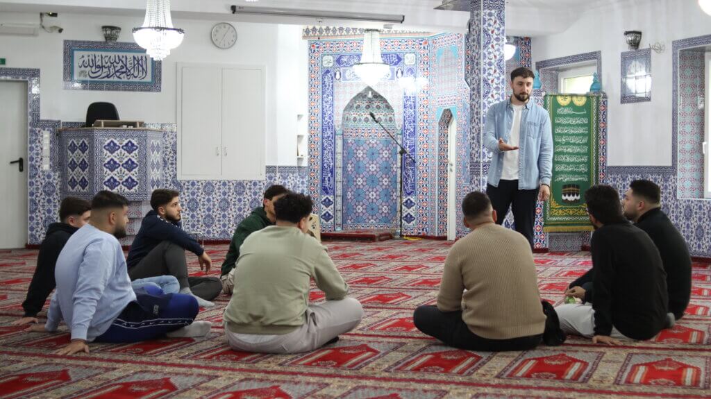 7 junge Männer sitzen mit dem Rücken zum Betrachter auf dem Boden im Halbkreis vor einem stehenden Referenten in einer Moschee