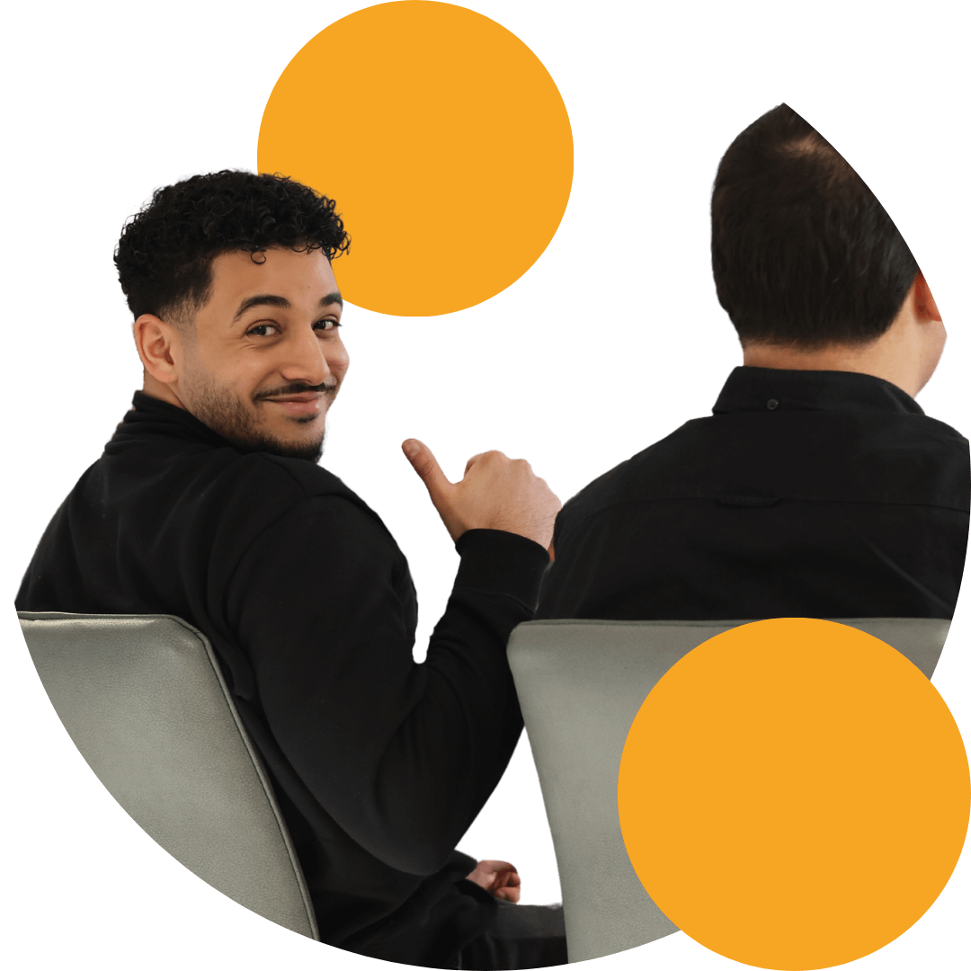 Ein Mann mit lockigen schwarzen Haaren und einem schwarzen Pullover sitzt auf einem Stuhl und schaut über die Schulter mit einem freundlichen Lächeln, während er einen Daumen hoch zeigt. Neben ihm eine andere Person, die mit dem Rücken zur Kamera sitzt. Die Szene ist grafisch gestaltet mit zwei großen, orangefarbenen Kreisen im Hintergrund.