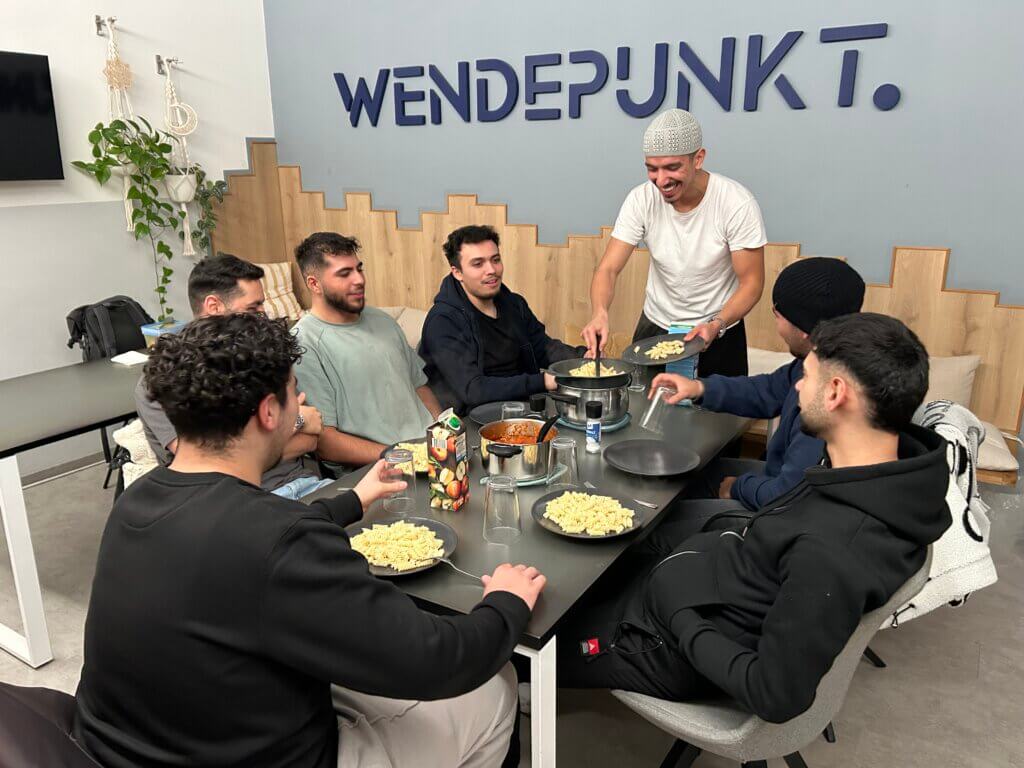 Sieben junge Männer sitzen um einen gedeckten Tisch. Einer löffelt Nudeln auf einen Teller. Im Hintergrund steht auf der Wand Wendepunkt geschrieben