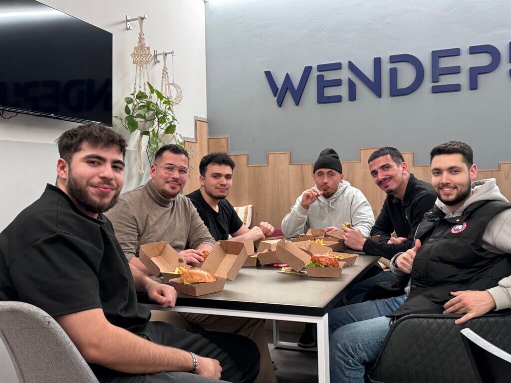 Sechs junge Männer sitzen um einen Tisch auf dem Essen steht. Sie lachen in die Kamera.