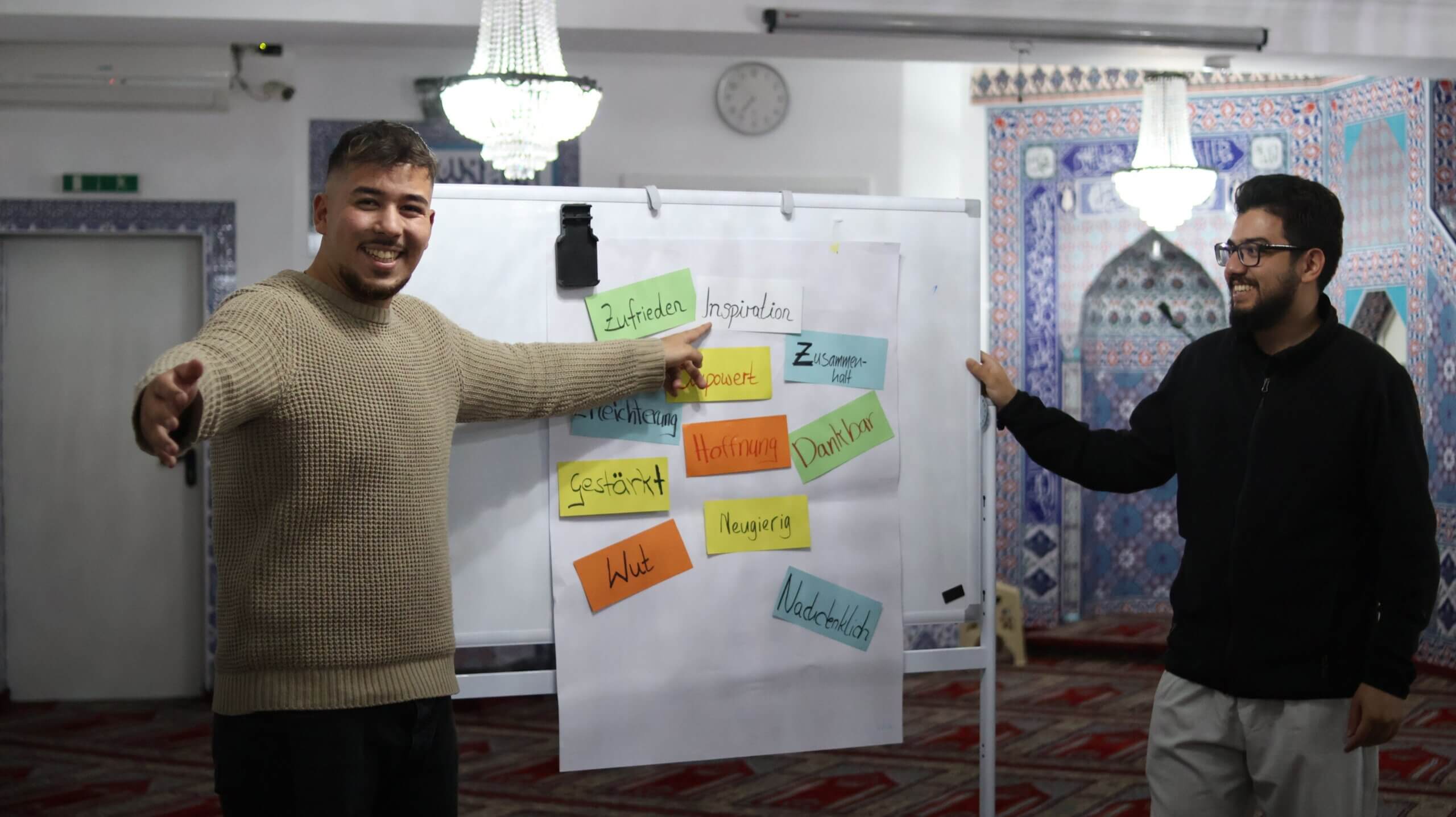 Zwei junge Männer stehen freudestrahlend links und rechts neben einer Plipchart mit bunten positiven Schlagwörtern darauf.