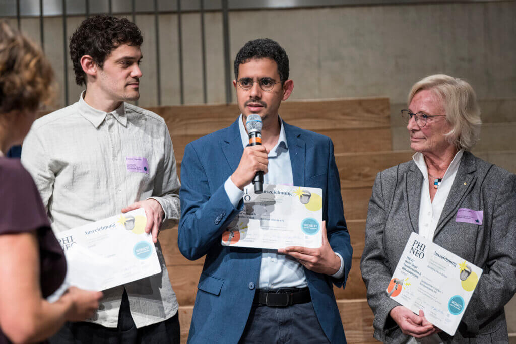 Drei Personen mit dem Phineo-Wirkt-Siegel in der Hand. In der Mitte spricht Mimoun Berrissoun in ein Mikrophon.